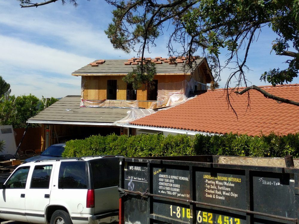 House under construction with exposed framing, new roof tiles, and a dumpster in front.