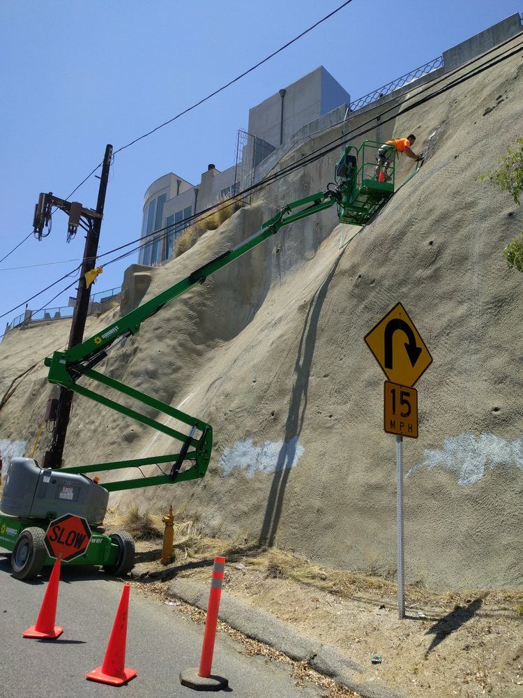 A worker in an aerial lift trims a hillside near a road; power lines and a large house are above.