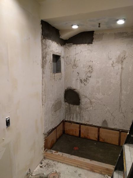 Bathroom under construction, cement walls with a niche, wooden panels at the base, and a tiled shower floor.