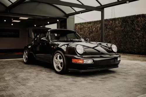 Black Porsche 911 parked under a canopy in a driveway, front-left view