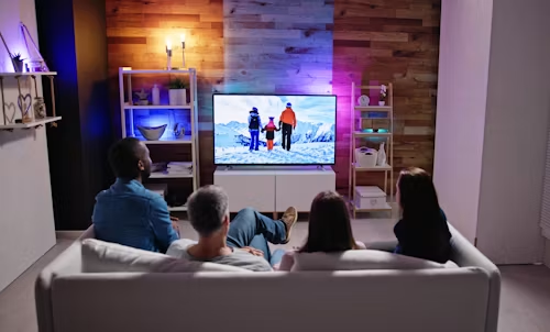 People on a couch watching TV in a cozy living room with a lit fireplace and warm wood accent wall