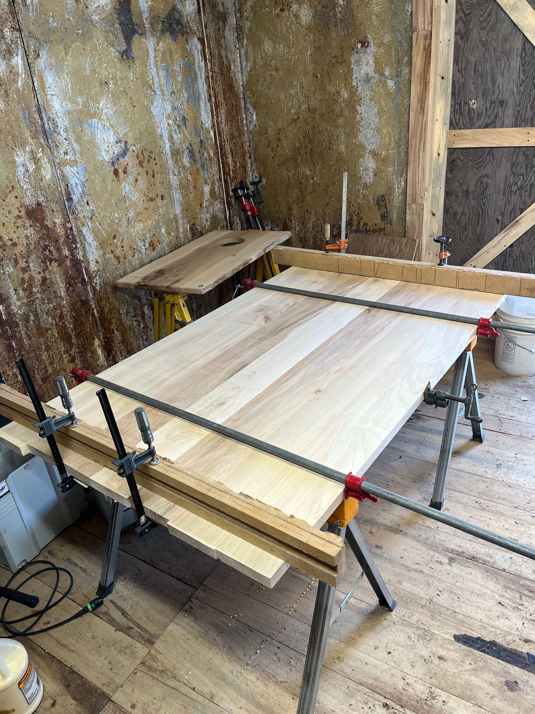 Wooden tabletop being glued, clamped on a sawhorse in a rustic workshop.