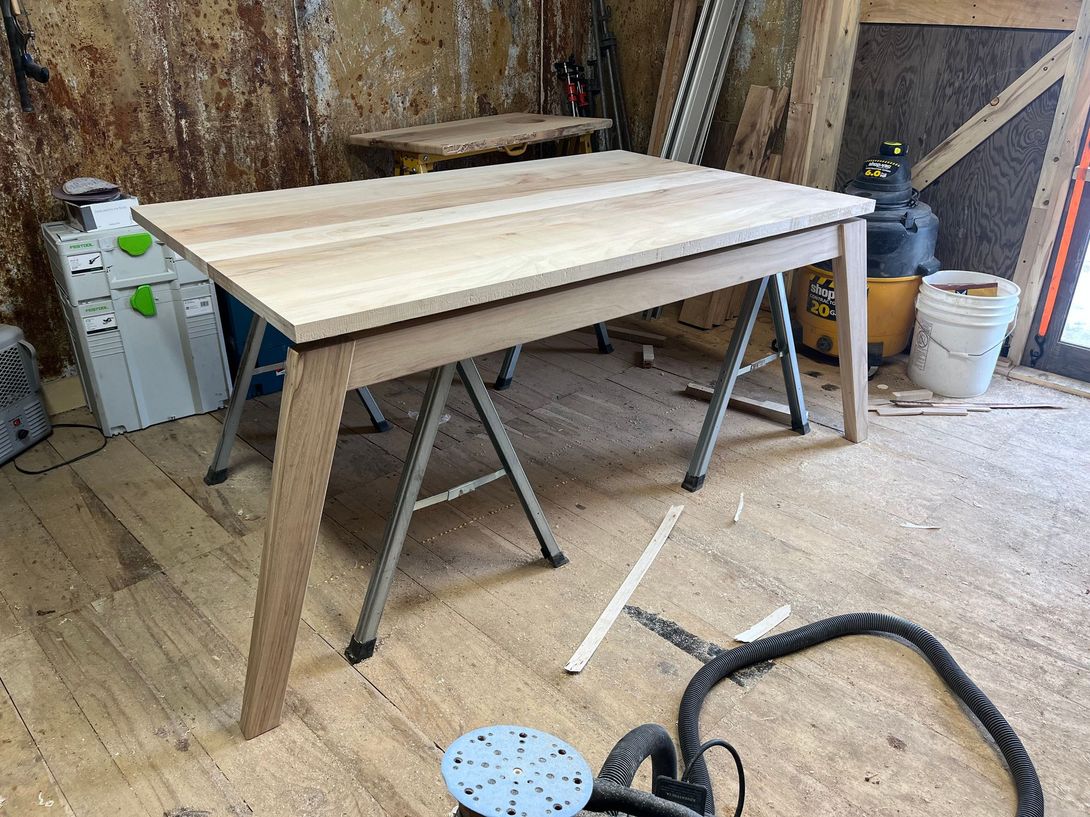 Wooden table under construction in a workshop, resting on metal sawhorses, tools and sawdust visible.