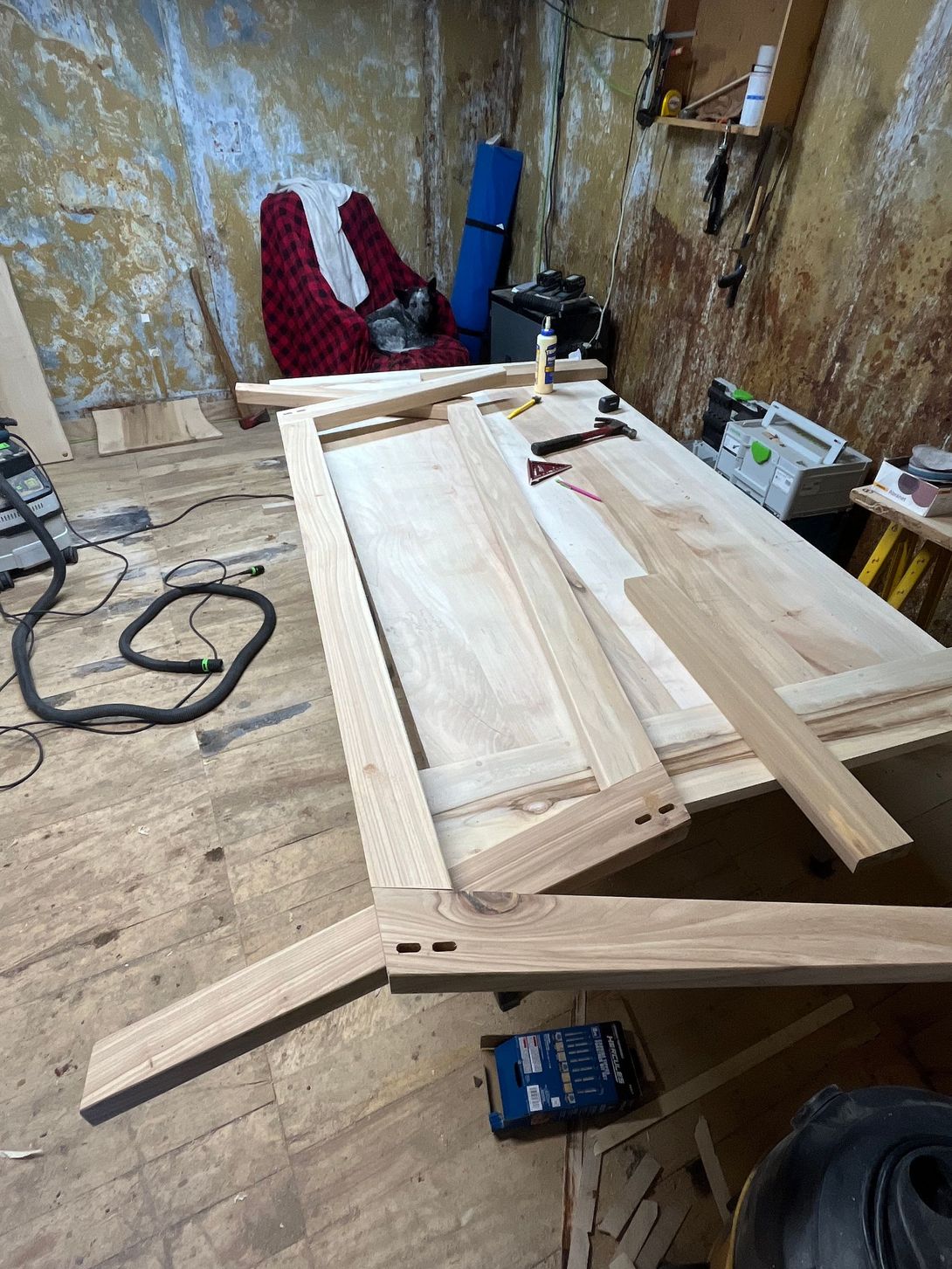 Woodworking shop with wooden door frame construction underway. Light wood, tools scattered on the floor, messy workshop setting.