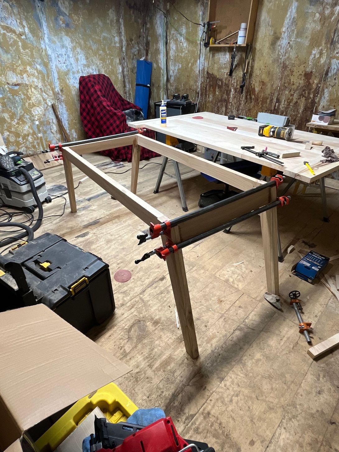 Wooden table frame being built in a cluttered workshop, with clamps and tools.