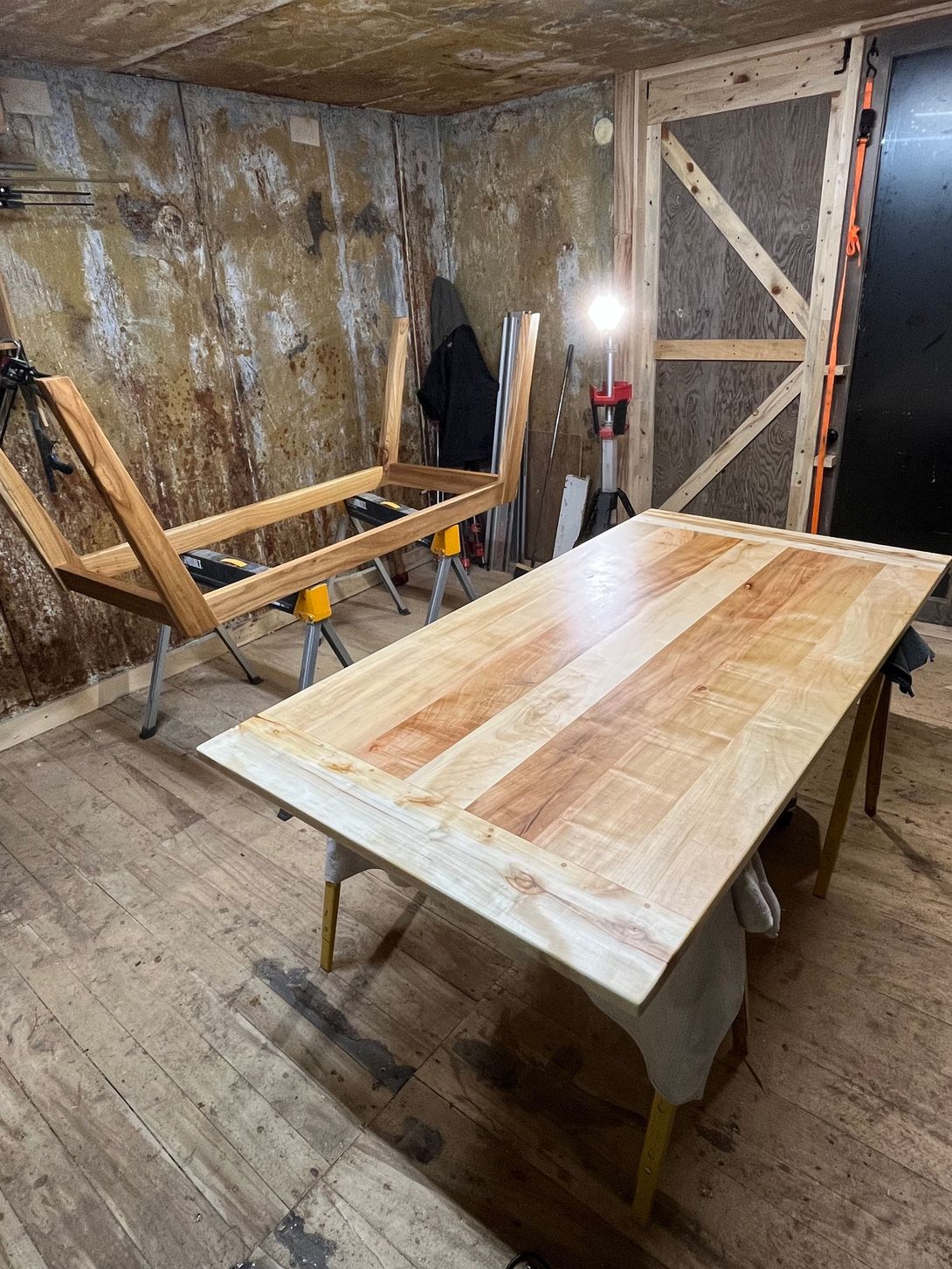 Wooden table being built in a workshop; unfinished frame, sawhorse, and spotlight in view.