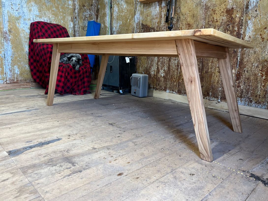 Wooden table with angled legs in a rustic room; a dog lounges on a nearby red chair.