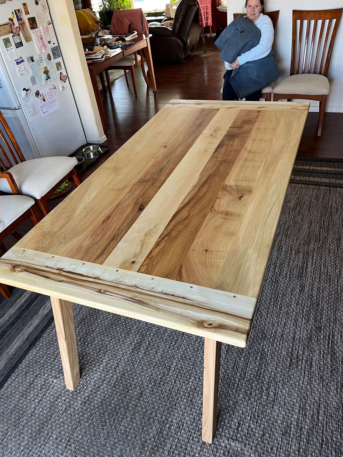 Wooden dining table with natural wood tones, a woman in the background.
