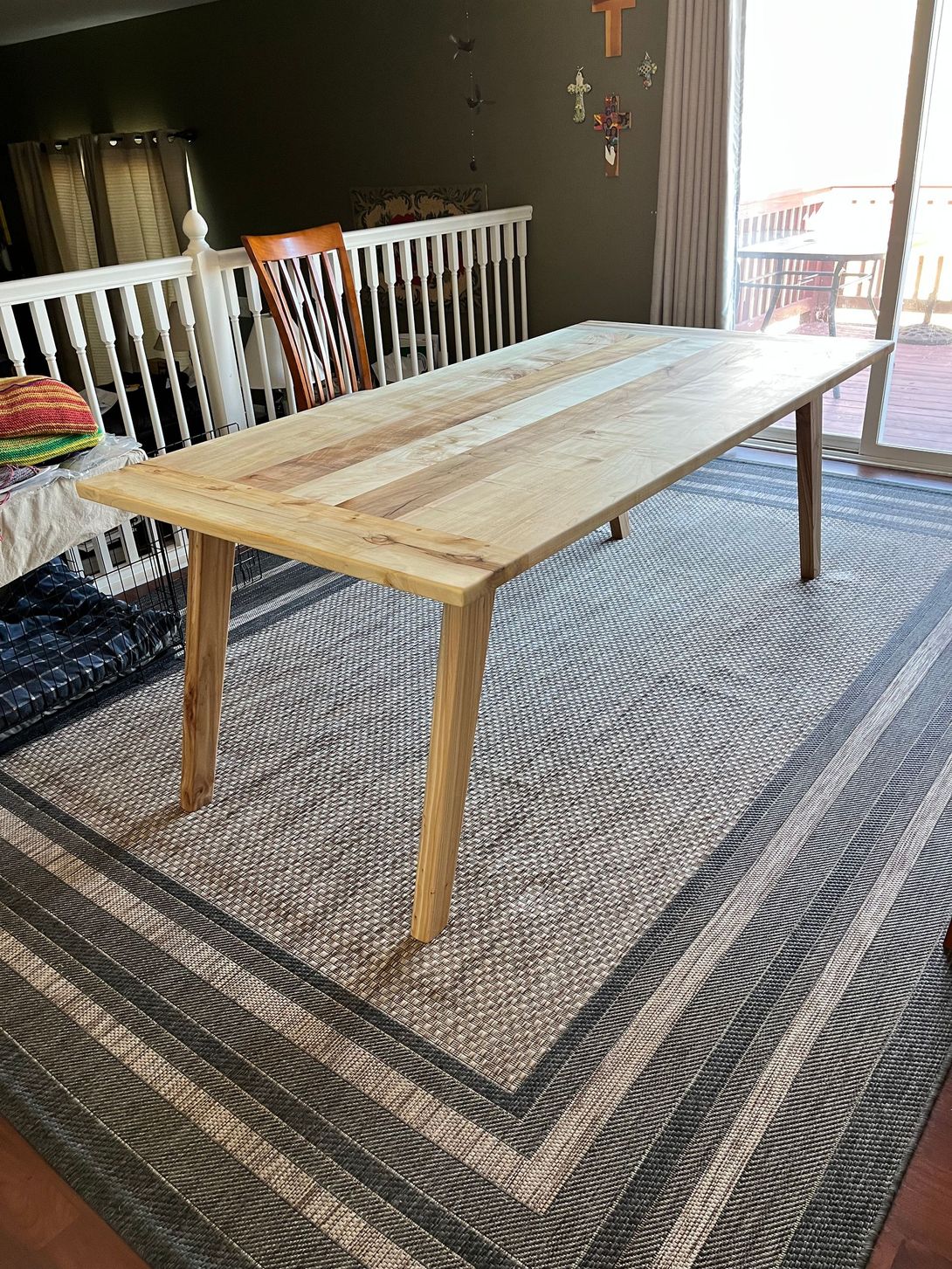 Wooden dining table on patterned rug, in a bright room, with white railing and a sliding glass door.