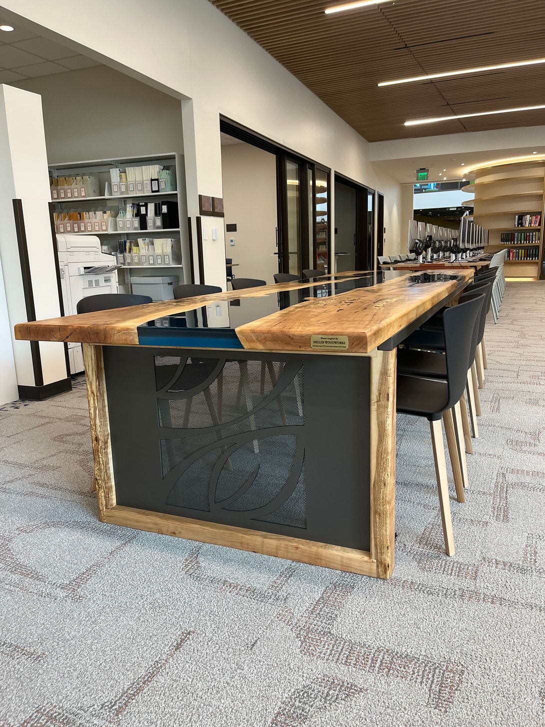 Long wooden table with chairs in a library setting. Dark gray metal base and wood trim.