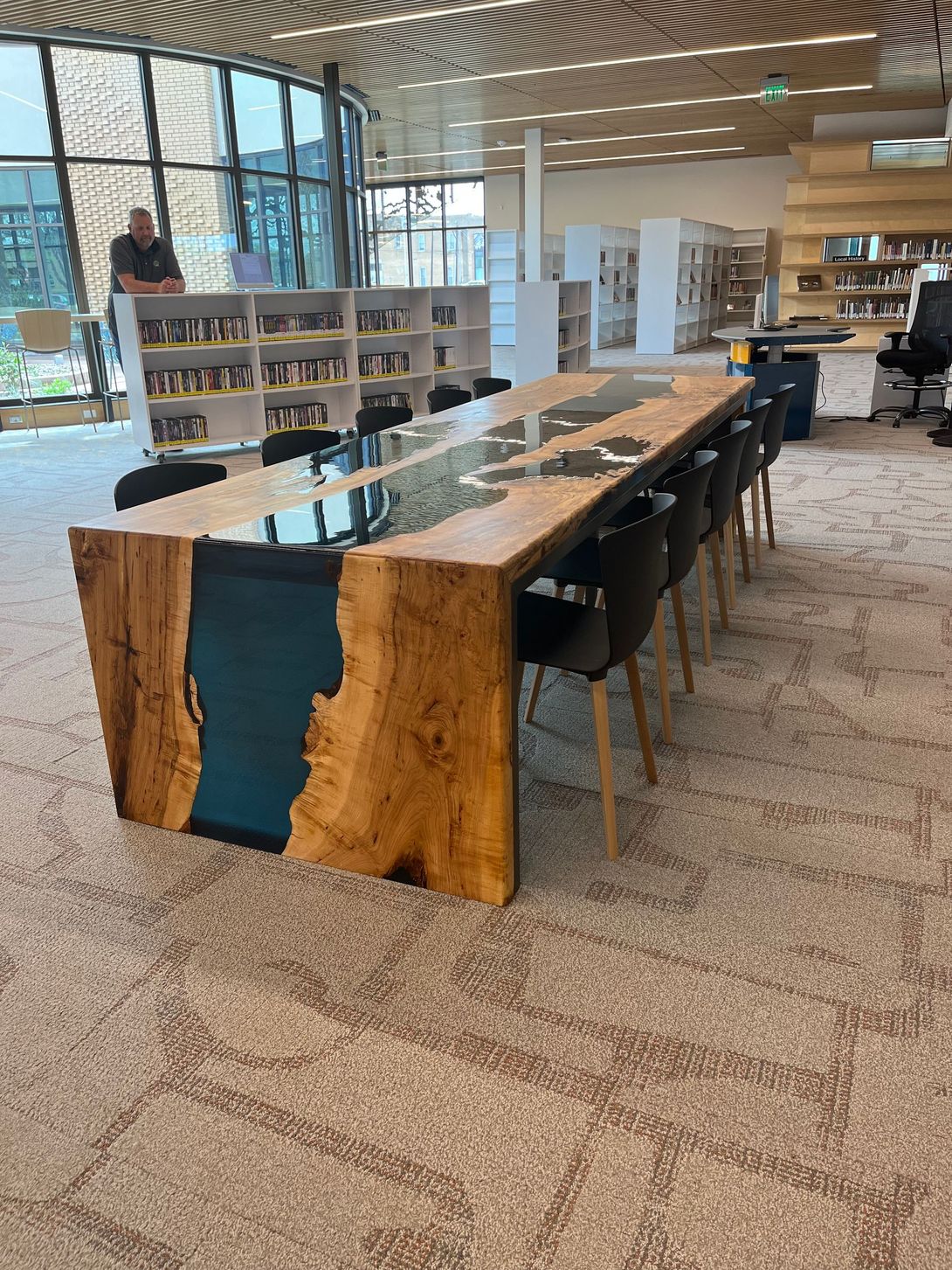 Long wooden table with blue resin inlay, black chairs in a modern library.