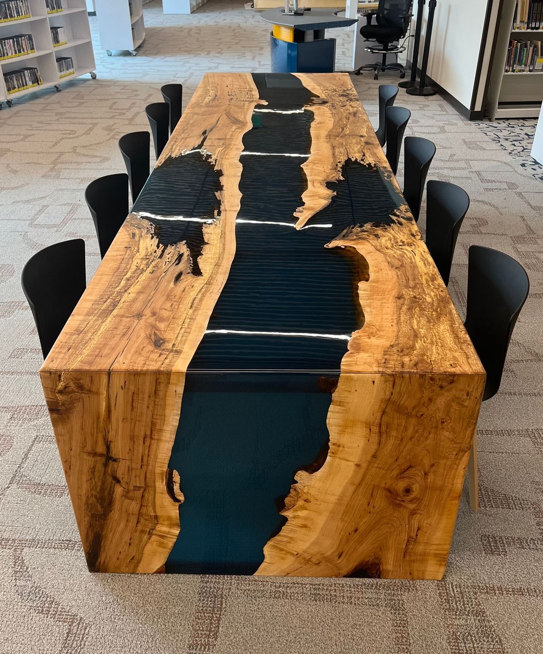 Wooden table with blue epoxy river, surrounded by black chairs, in a library setting.