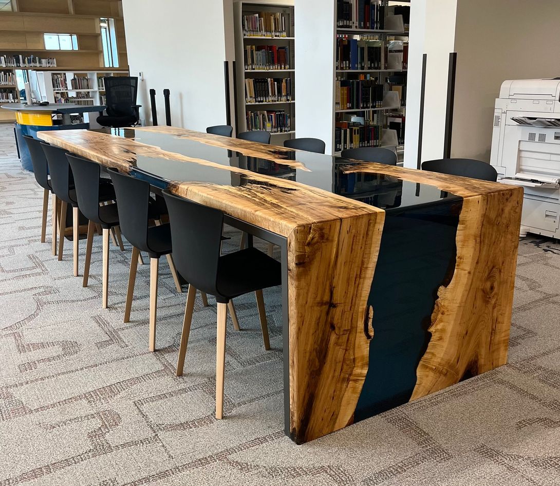 Large wooden table with dark blue epoxy and black chairs in a library setting.