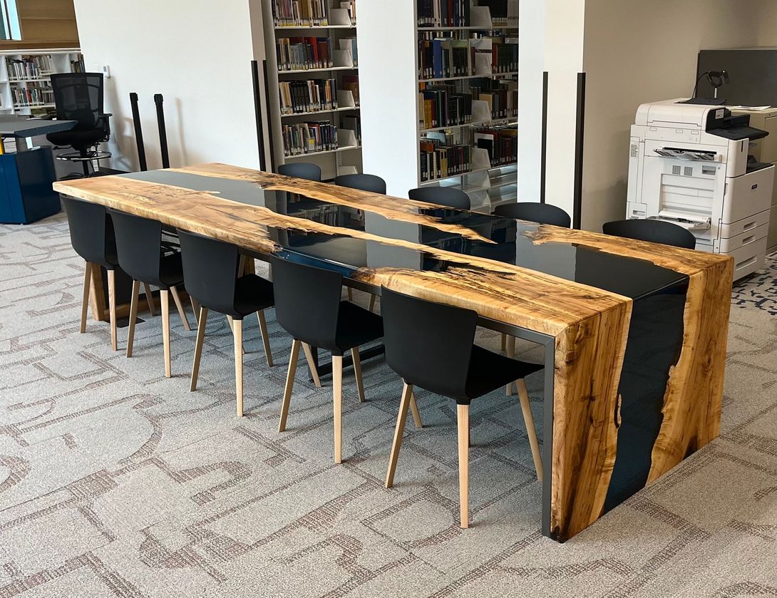 Long wooden table with black resin center, black chairs, library setting.