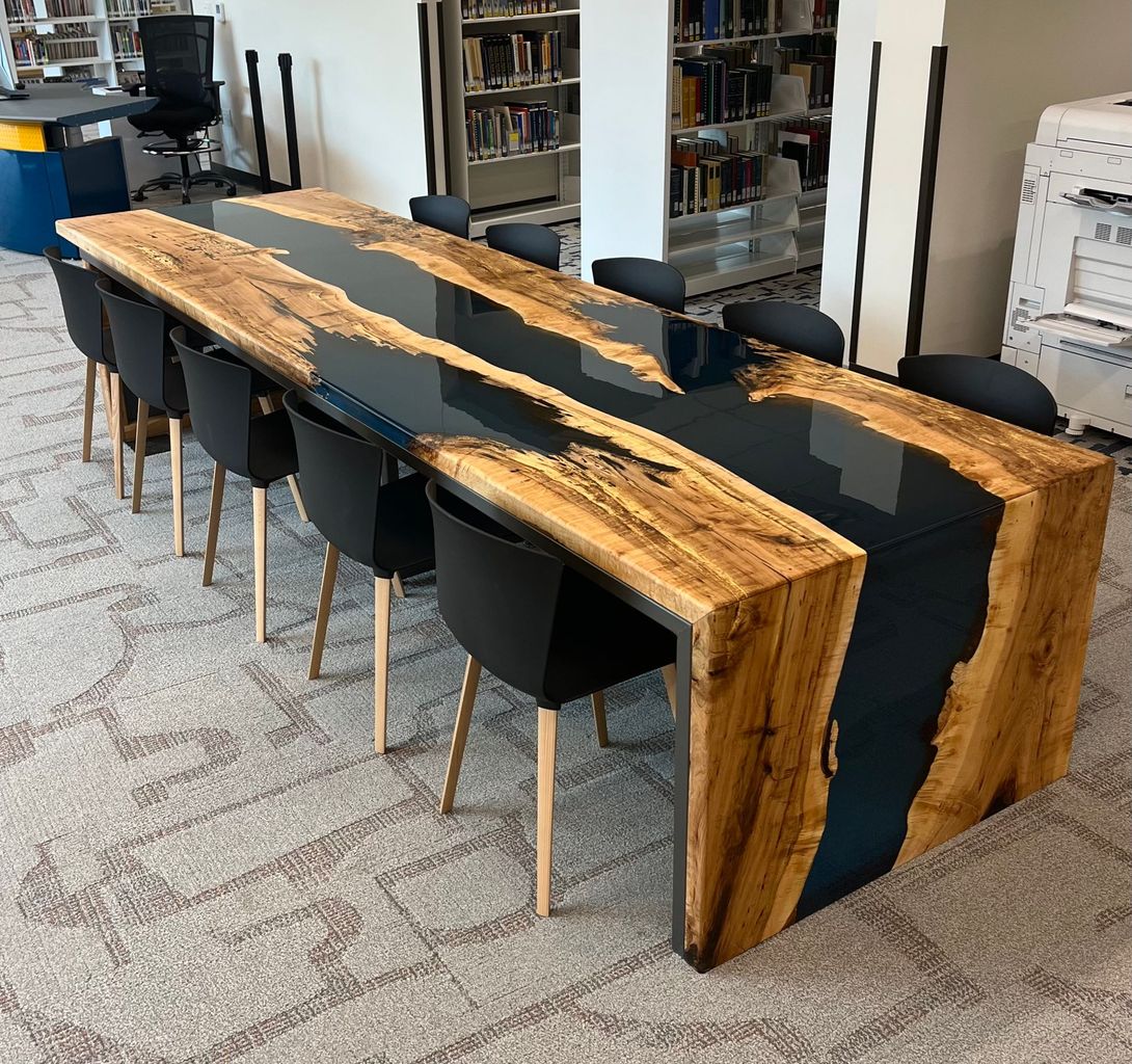 Long wooden table with black resin center, surrounded by black chairs, in a library.