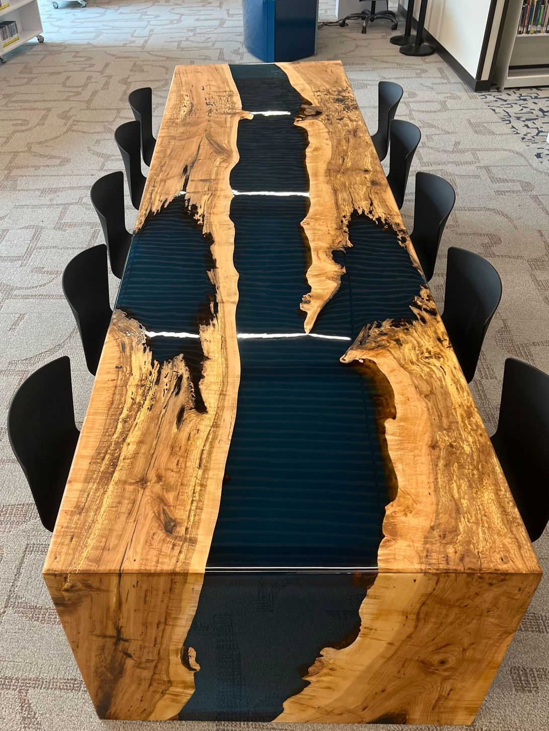 A large wooden table with a blue epoxy river, surrounded by black chairs, in a library setting.