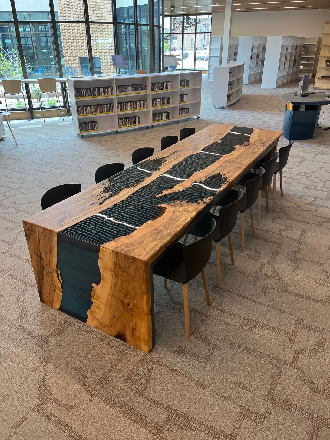 Large wooden table with black resin inlay, surrounded by black chairs, in a library.