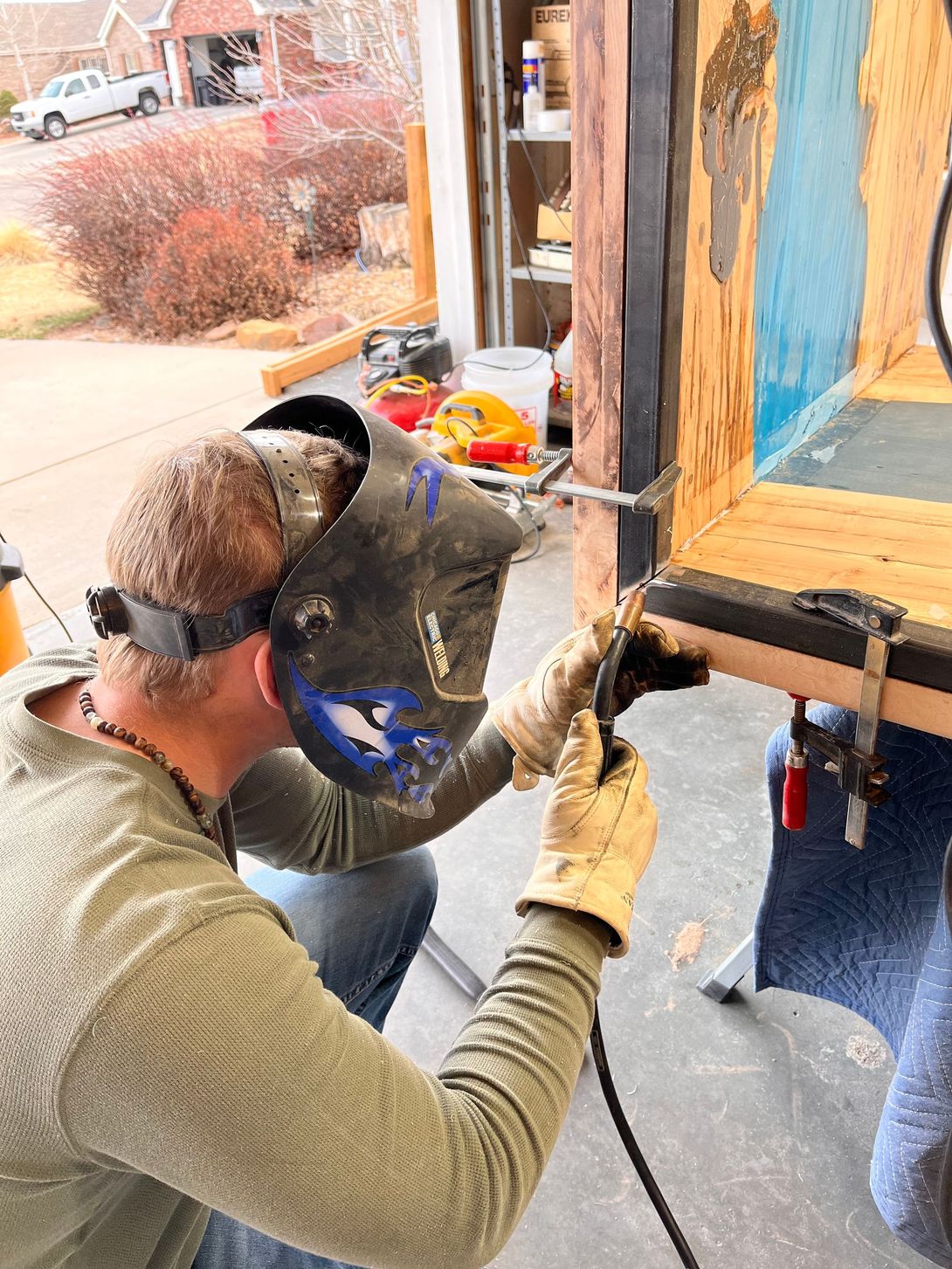 Man welding metal in a garage; wearing a welding mask and gloves. There's a house and car visible outside.