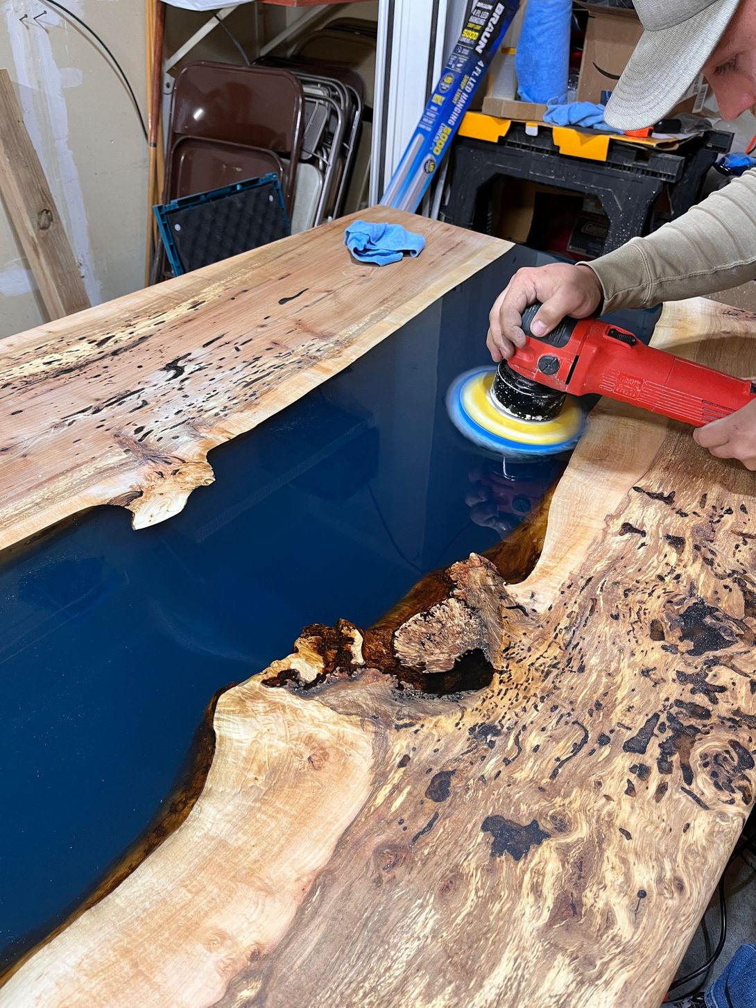Man using a buffer on a blue epoxy river table, in a garage workshop.
