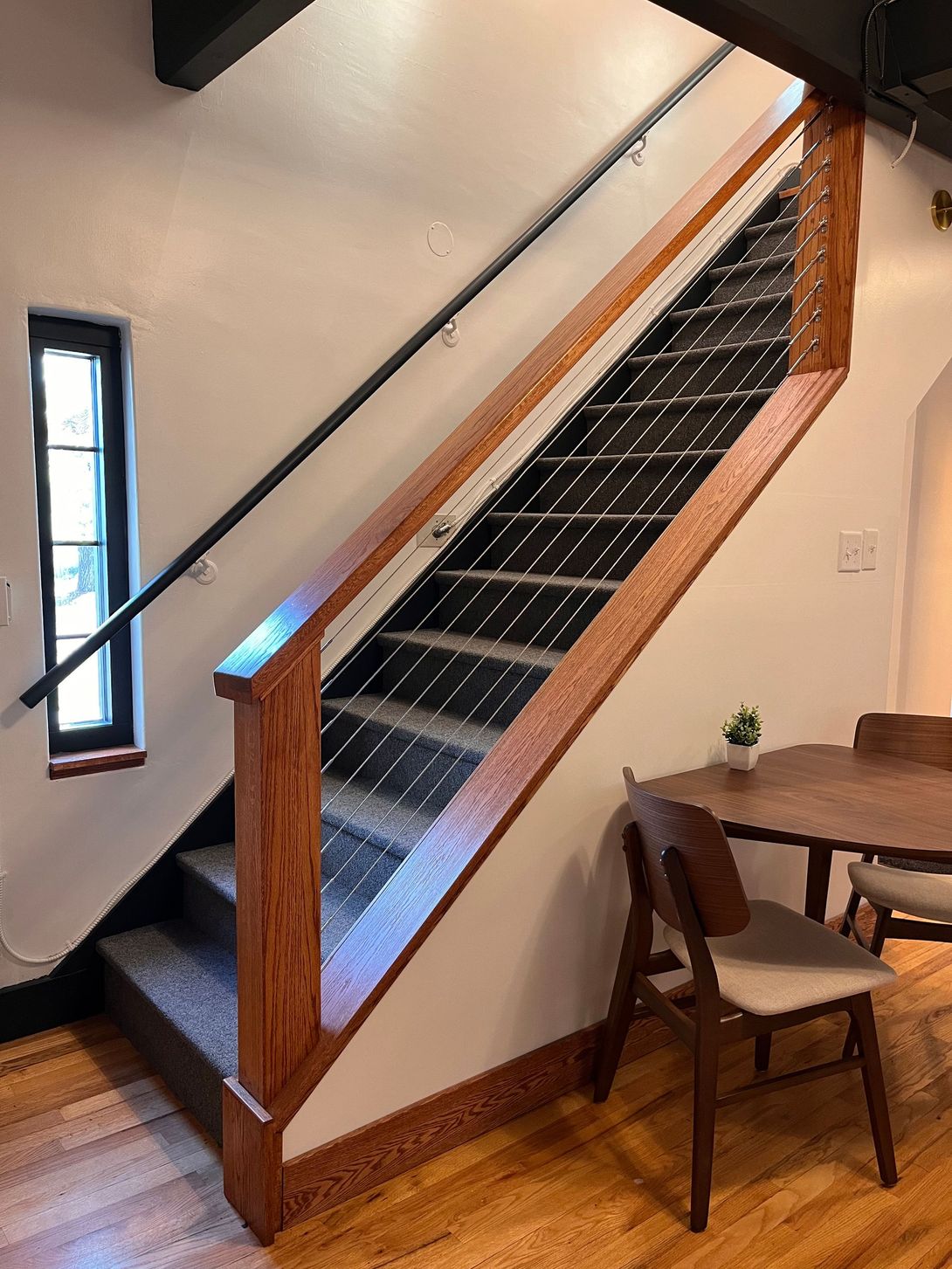 Staircase with wood railing, steps, and a black handrail against a white wall. Dining table and chairs are visible.