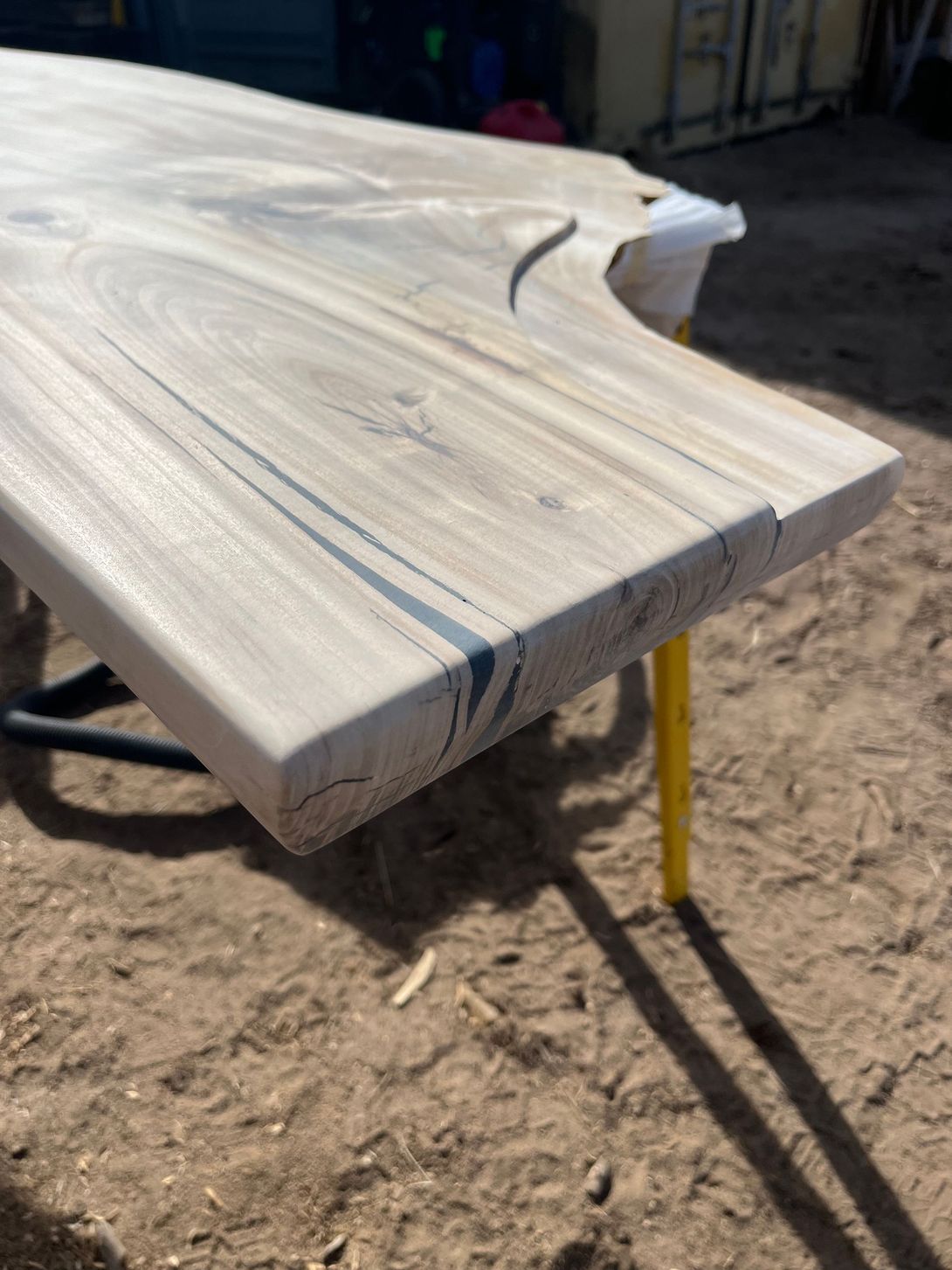 Wooden table, light brown with black streaks, resting on yellow and black legs outdoors on dirt.