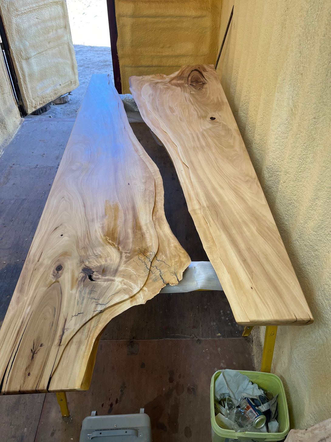 Two unfinished wood slabs, light brown, are set on yellow supports. One has a dark epoxy river.