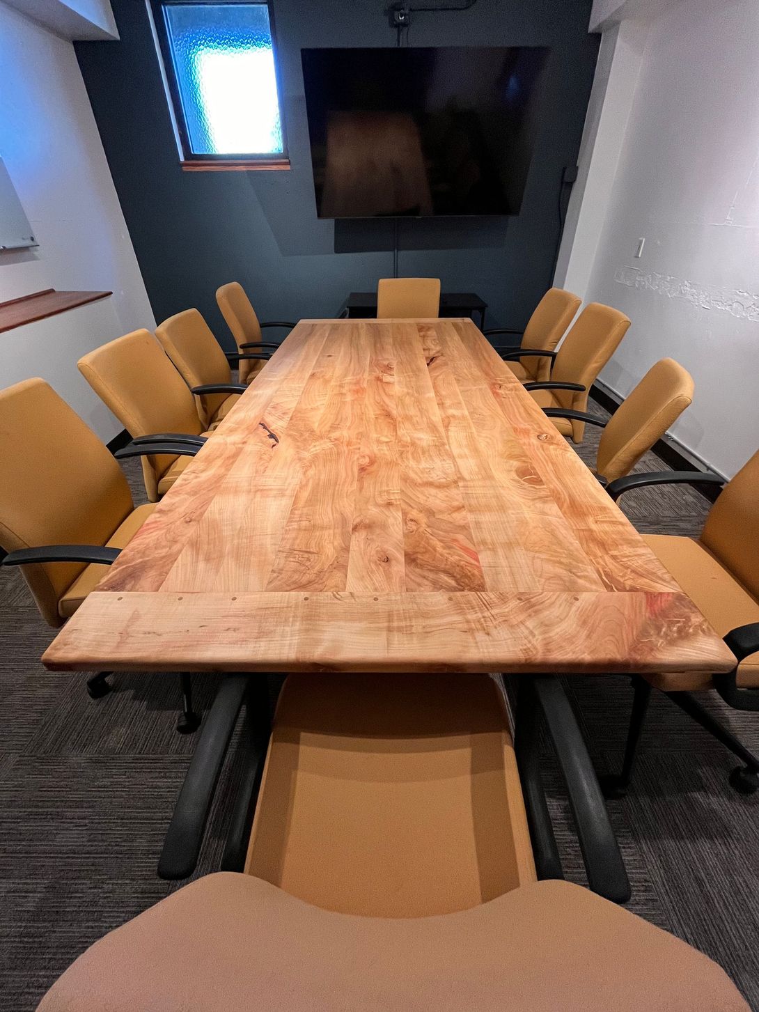 Conference room with long wooden table and tan chairs, dark wall with TV.