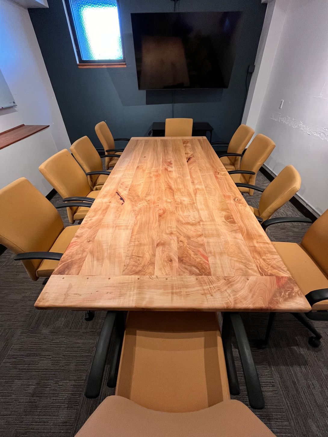 Conference room with a long wooden table and yellow chairs. A TV is mounted on the dark wall.