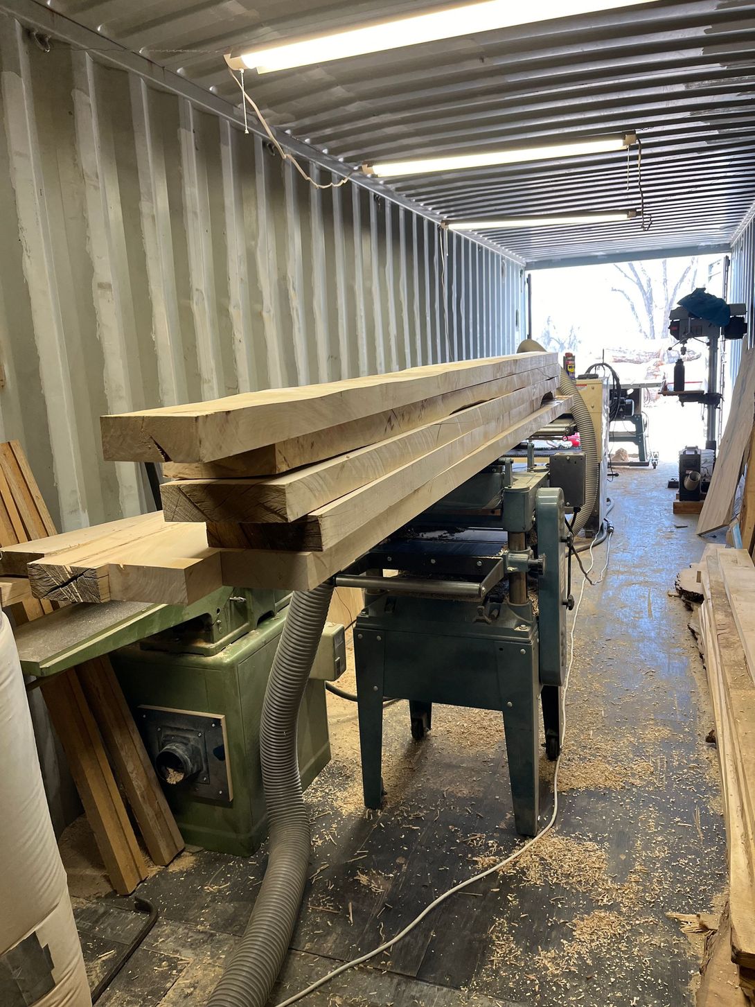 Wood planks on a planer in a workshop; container walls in the background.