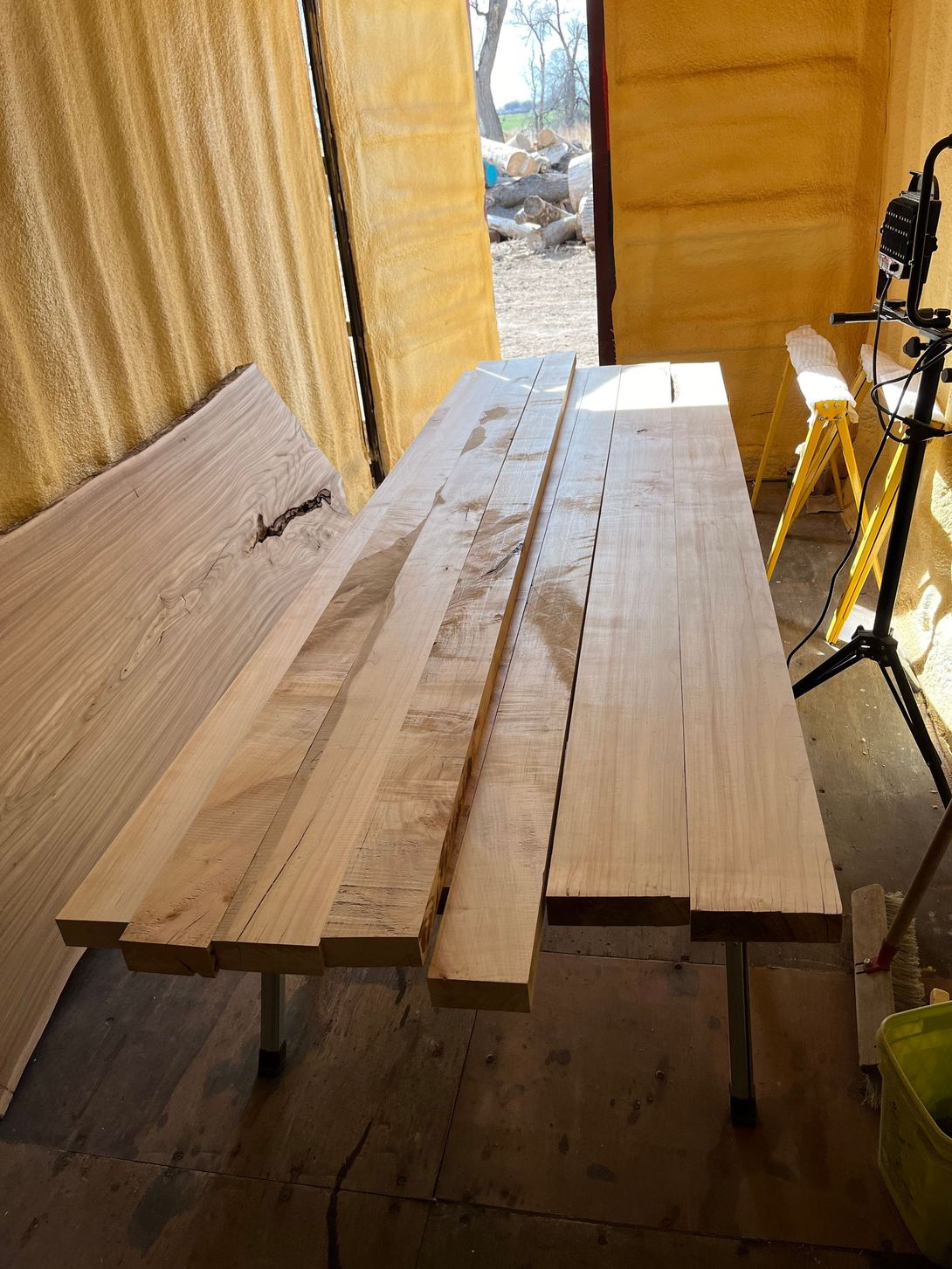 Wooden planks on a metal-legged table inside a room with corrugated walls; saw on a tripod to the right.