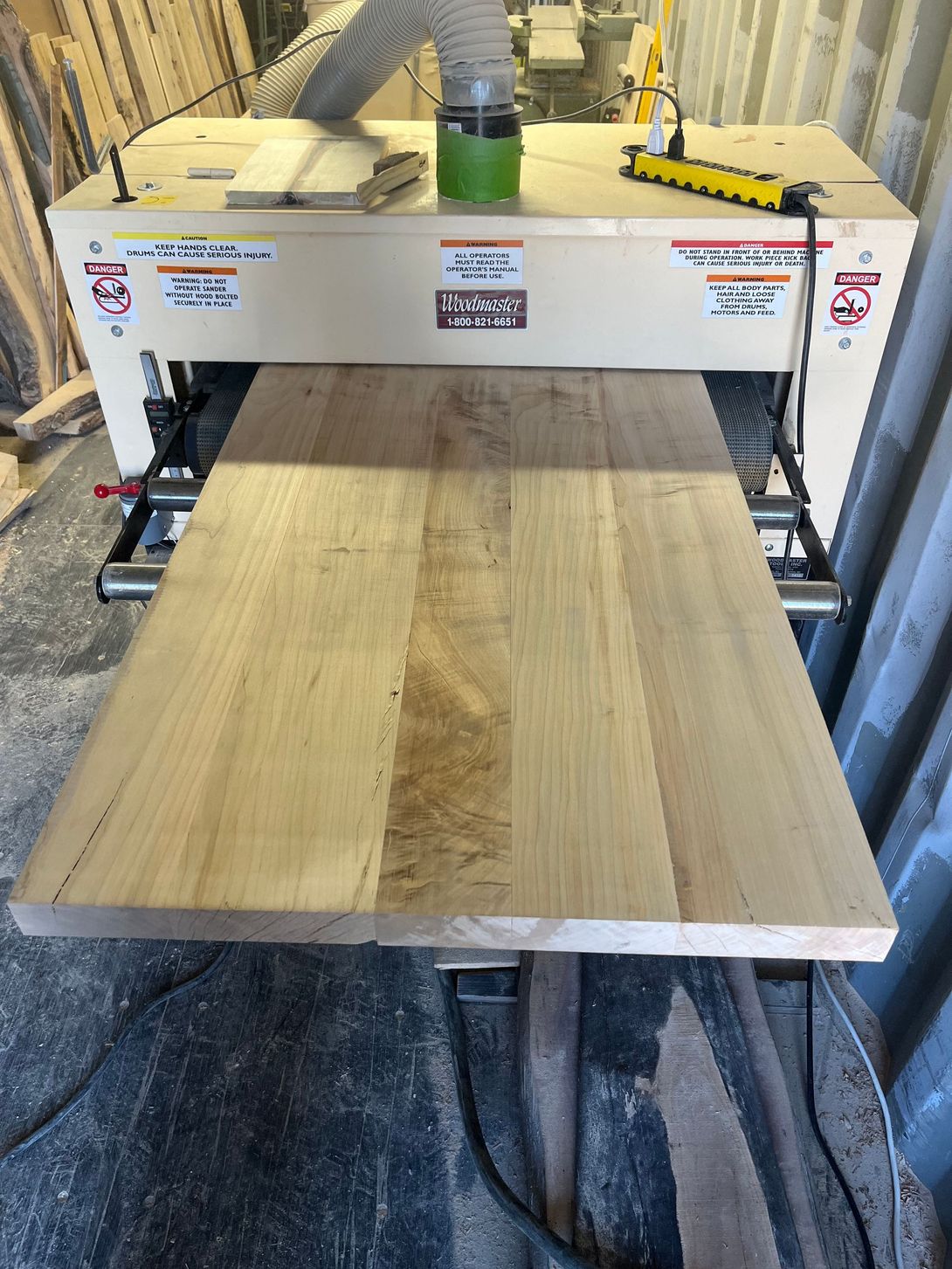 A large wooden tabletop being sanded by a wide-belt sander in a workshop.