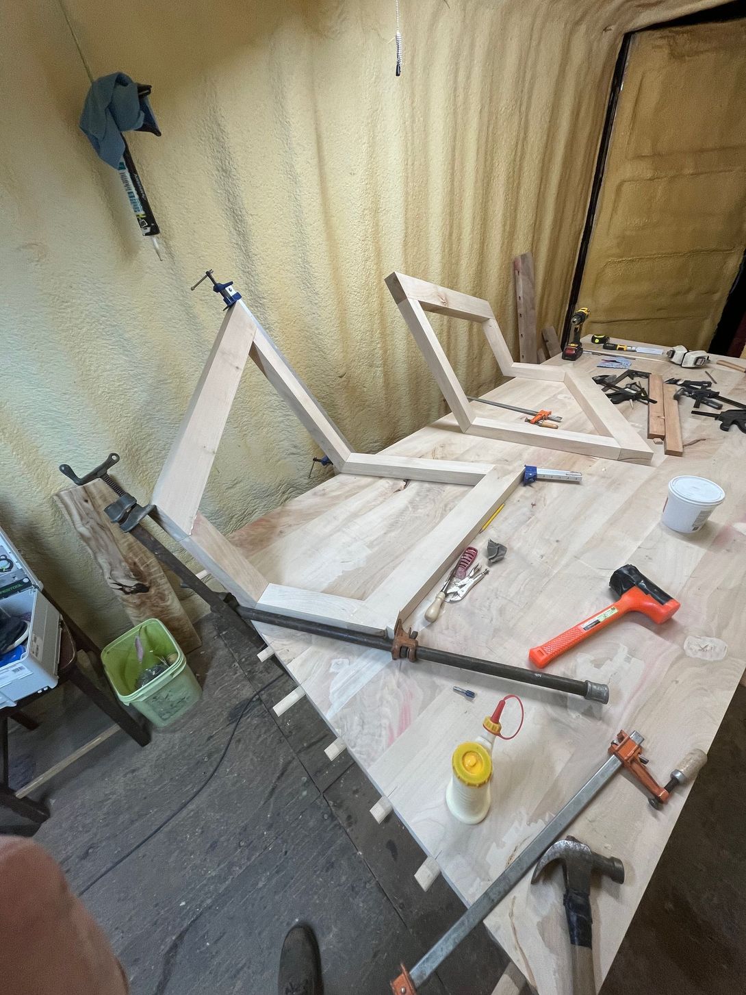 Woodworking scene: Two wooden chair frames clamped together on a work surface. Tools and supplies scattered nearby.