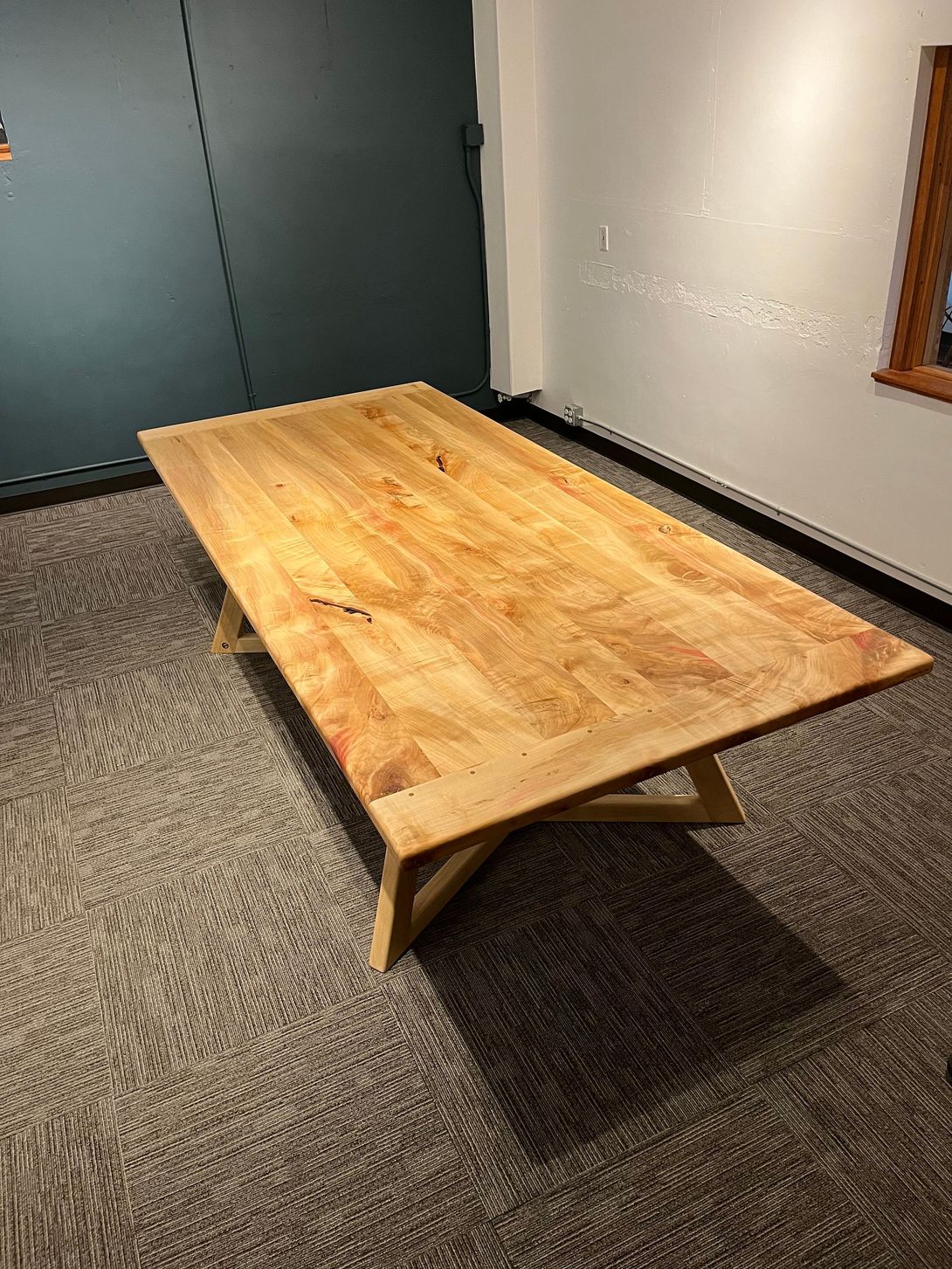 Wooden table with a light-colored top and angled legs, set on patterned carpet in a room with a blue and white wall.