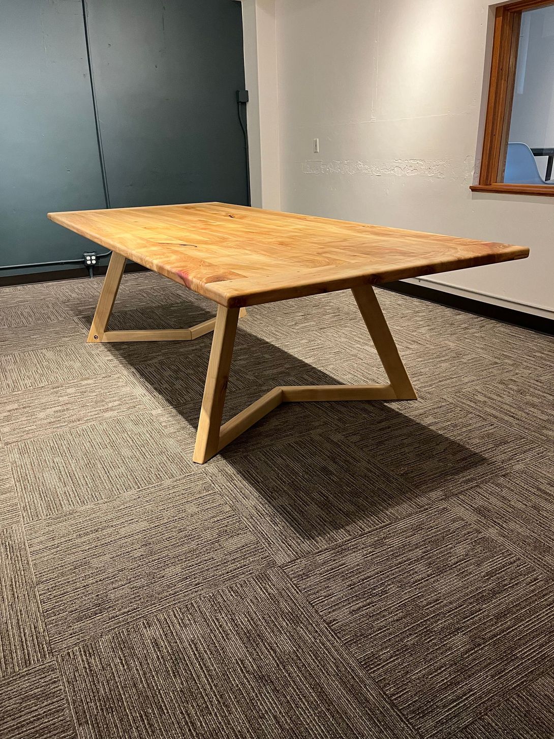 Wooden table with geometric legs on patterned carpet; room with neutral walls and a window.