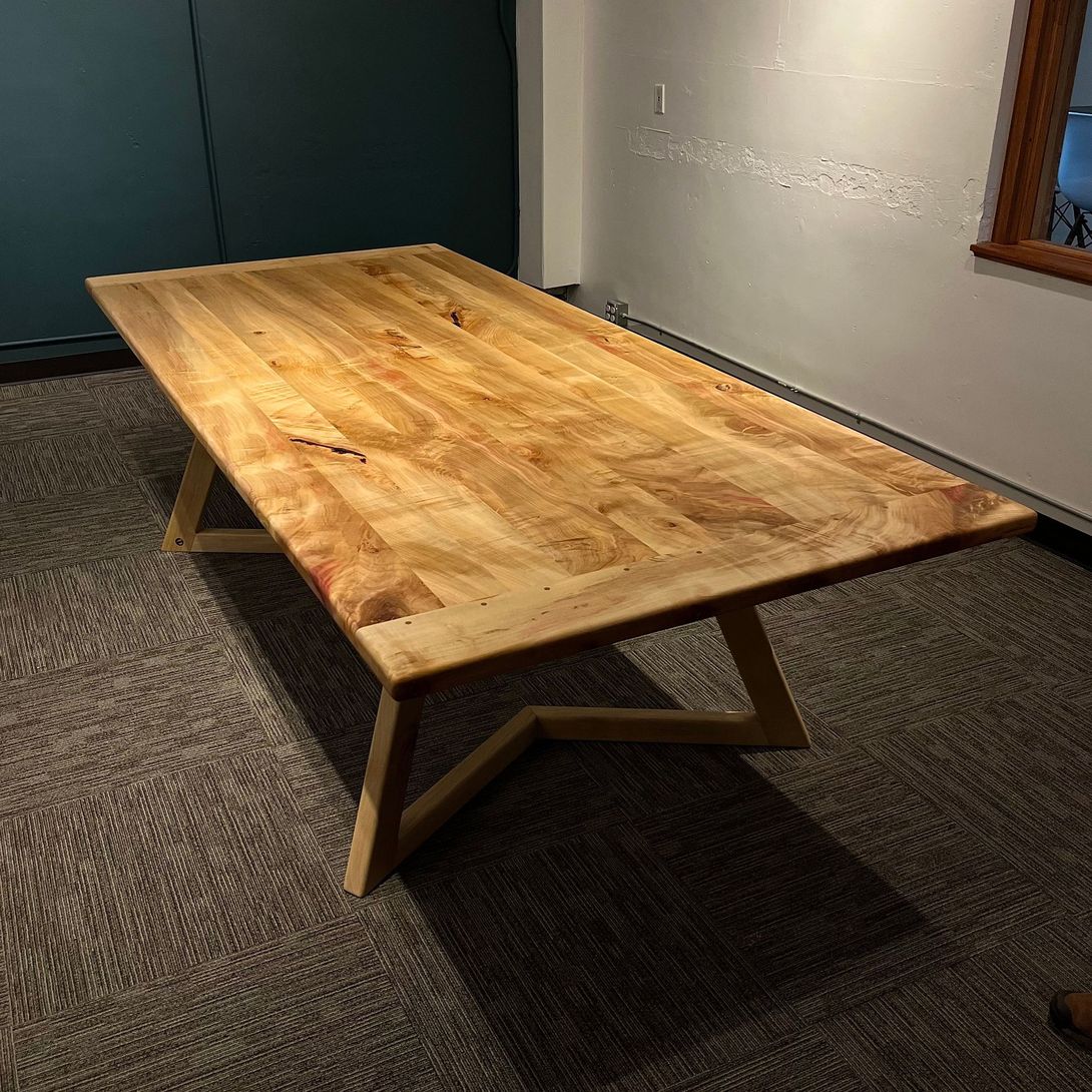Wooden rectangular dining table with light wood grain, angled legs, in a room with a carpeted floor.