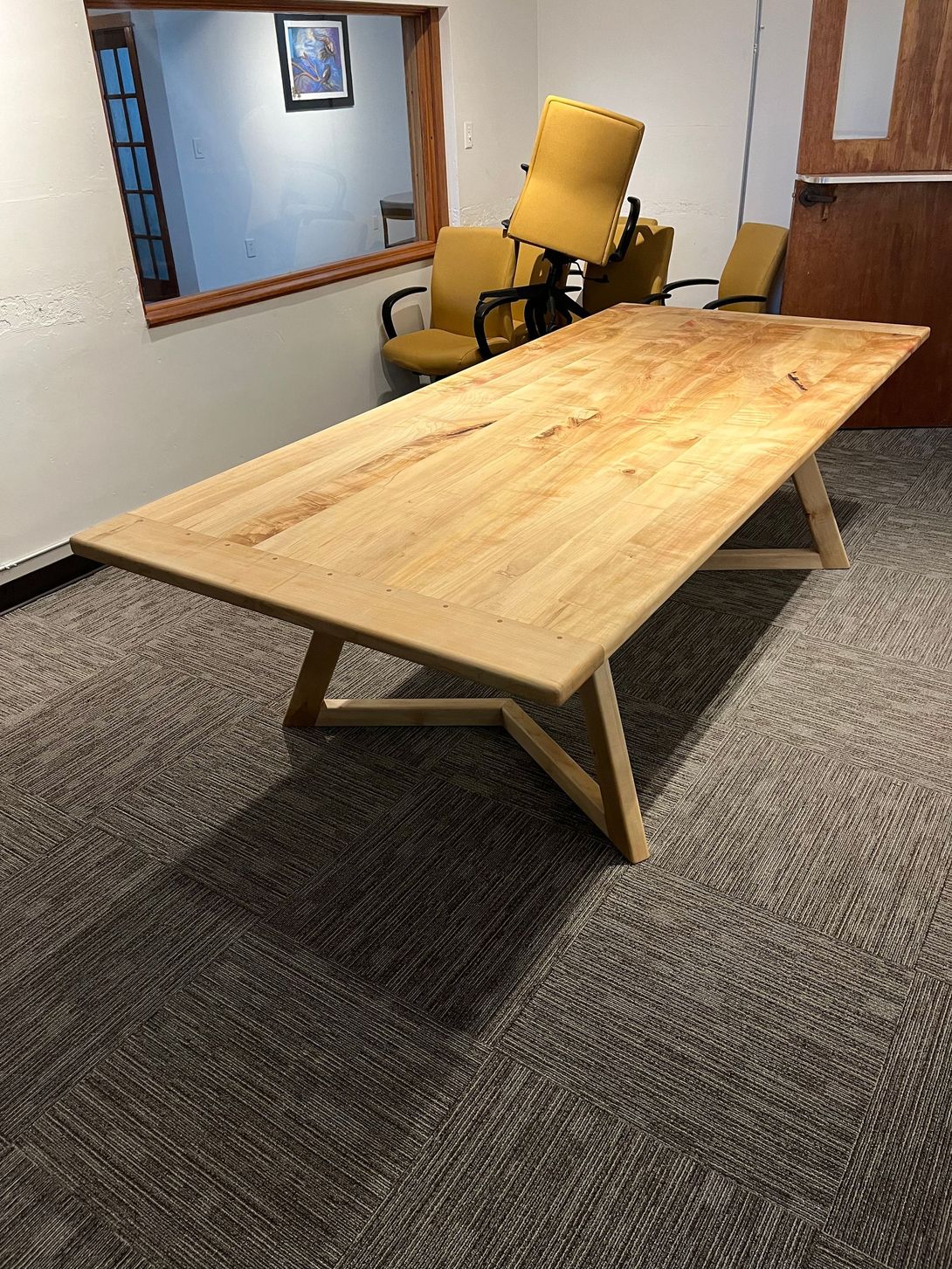 Light wood conference table on patterned carpet in a room, with yellow chairs and a window in the background.