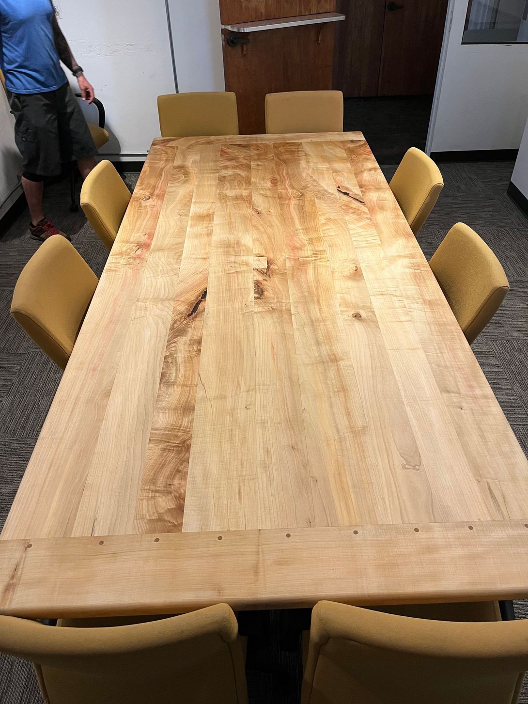A large wooden table with six yellow chairs. A person stands beside the table, holding a brush.