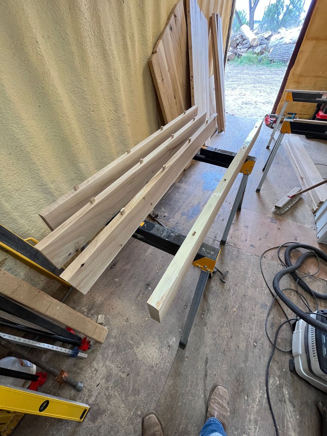 Wood planks on sawhorses in a workshop setting. Tools, sawdust, and wooden walls are also visible.
