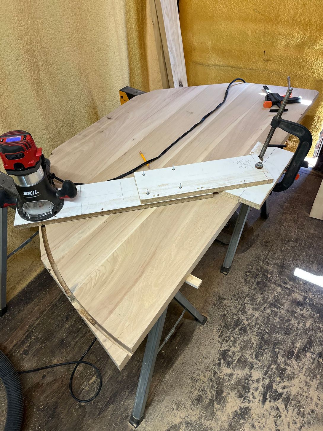 A router cutting a curved edge on a wooden table; a jig is clamped down to guide the tool.