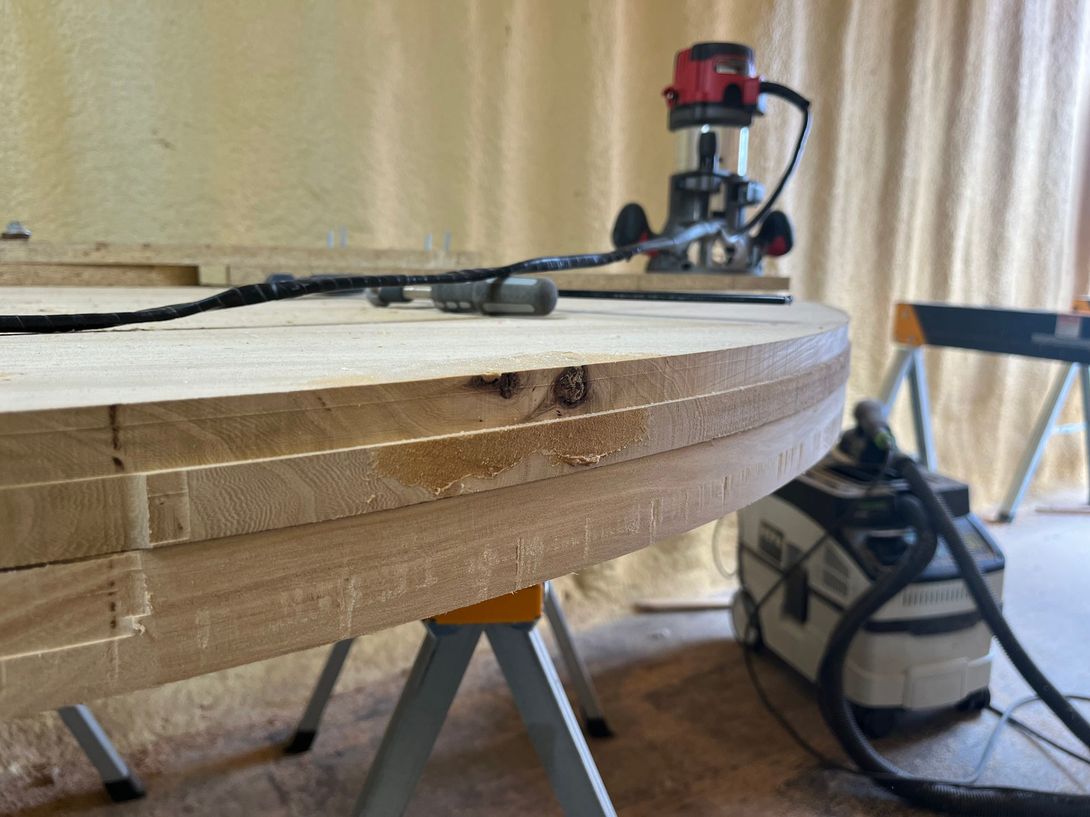 A router shaping the edge of a large, round wooden tabletop. Workshop setting, light wood, sawhorse.