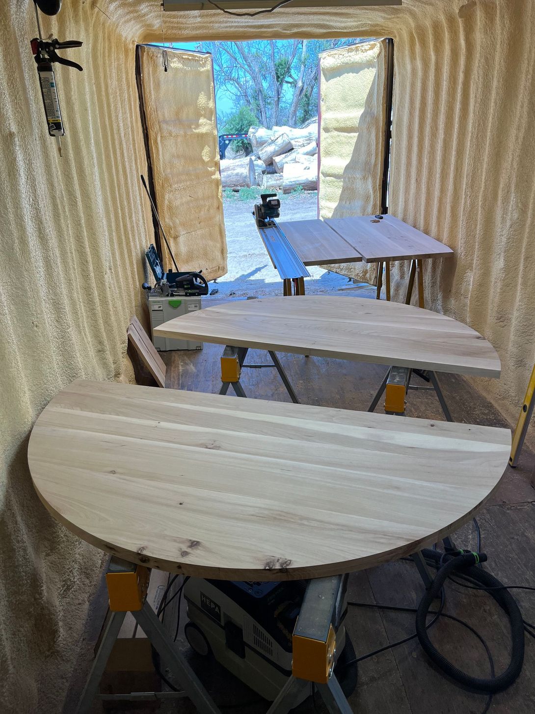 Two large, half-circle wooden tabletops on sawhorses inside a corrugated structure, open door in the background.