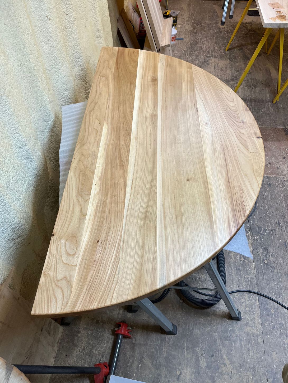 Half-circle wooden tabletop in a workshop. The unfinished wood has visible grain, resting on a sawhorse.