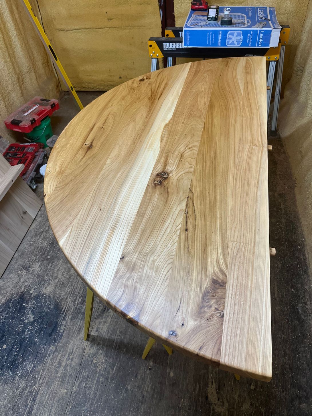 Half-circle wooden tabletop, light brown with wood grain, on yellow sawhorses in a workshop.
