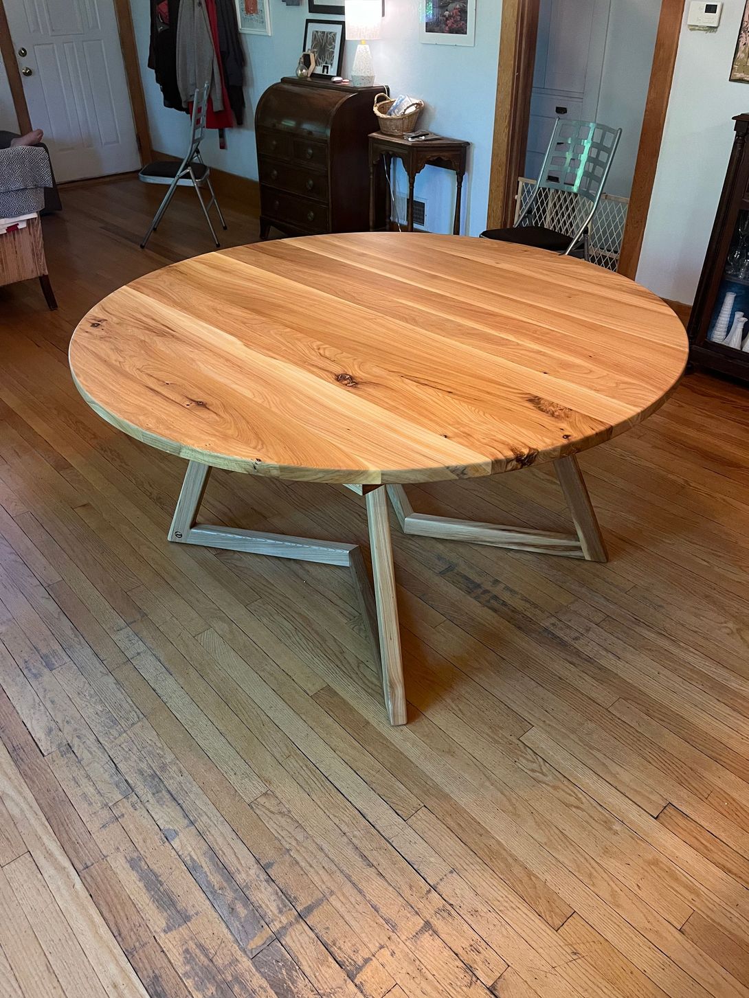 Round wooden table with a light-colored top and a geometric base, in a room with hardwood floors.