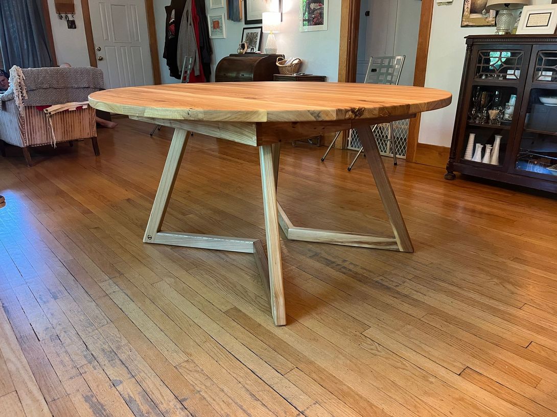 Round wooden dining table with angled trestle legs, on hardwood floor.