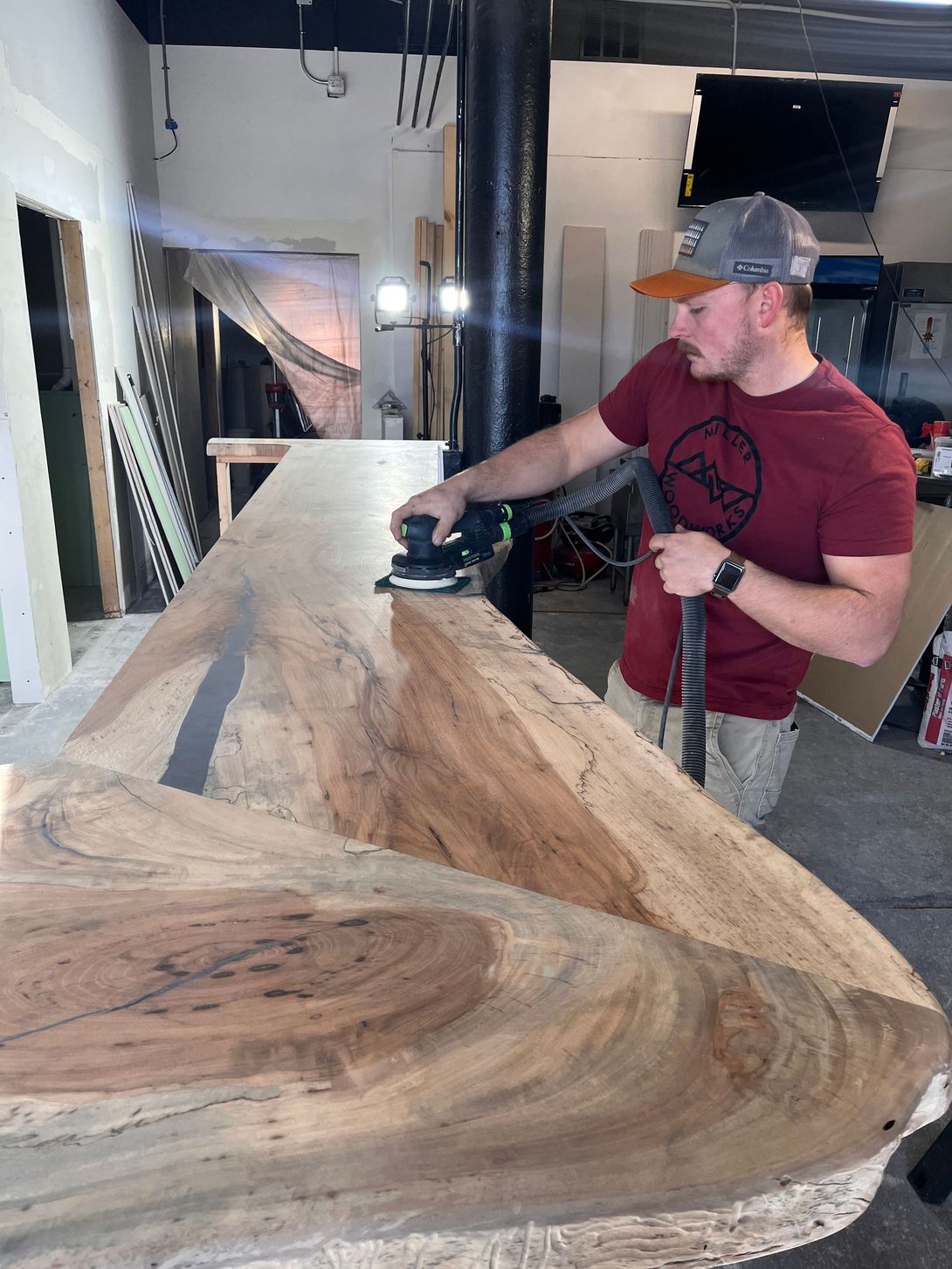 Man sanding a large wooden slab, wearing a red shirt and a cap, in a workshop.