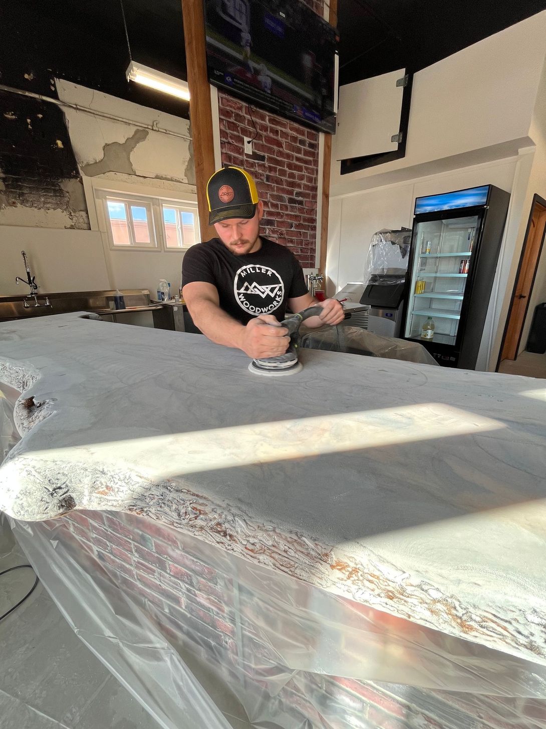 Man sanding a concrete bar top. He wears a hat and a black t-shirt in a construction setting.