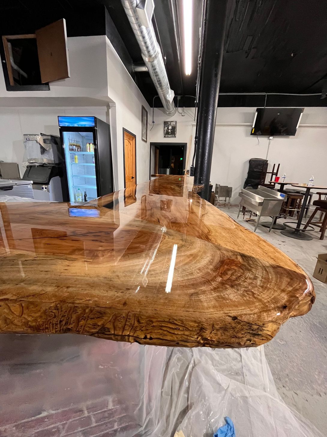 A bar with a glossy wooden countertop.  Drinks cooler, seating and doorway in the background.