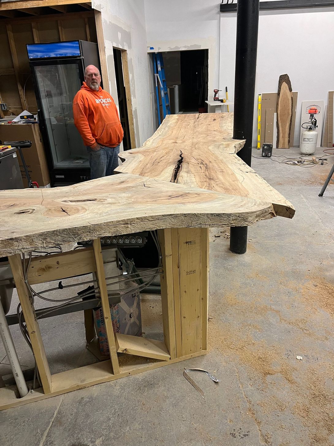 Man stands near a large, unfinished wooden bar in a workshop, wearing orange.
