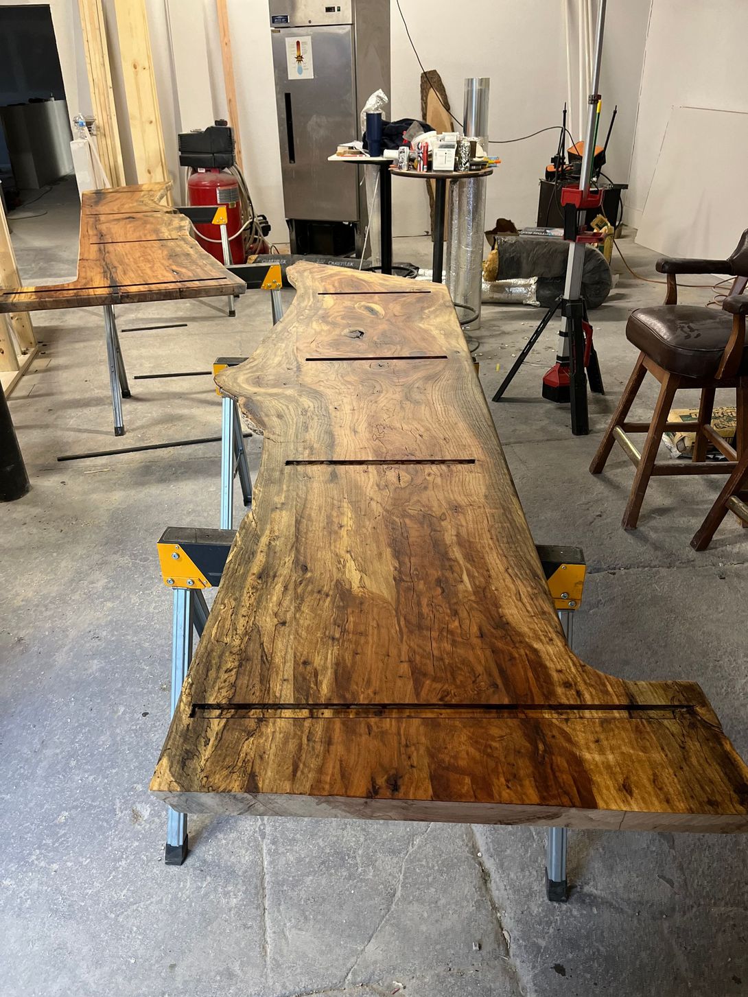 A long, wooden bar top with cut-out grooves sits on sawhorses in a workshop.